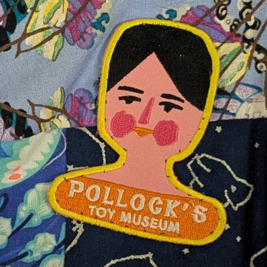 Sew-on patch featuring a Dutch Doll face and the text 'Pollock's Toy Museum', sewn onto a colourful patchwork background.